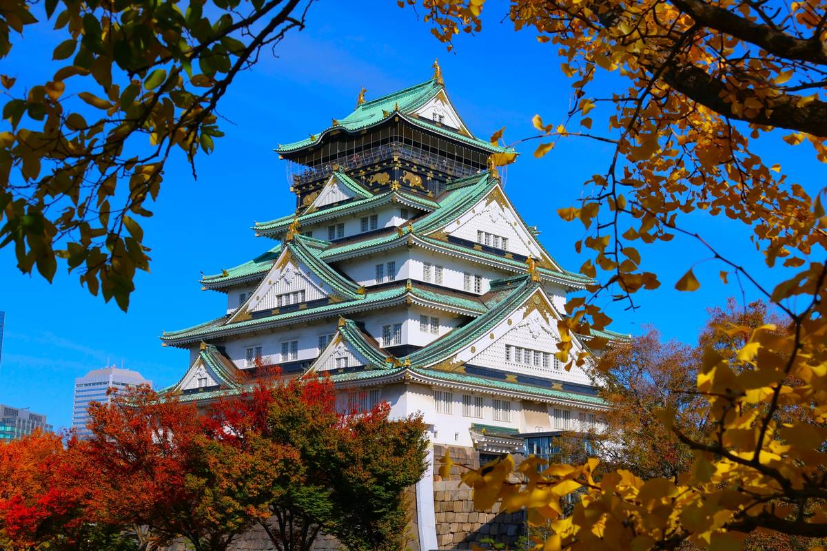 Osaka Castle with green roofs and white walls, framed by colorful autumn leaves against a blue sky.