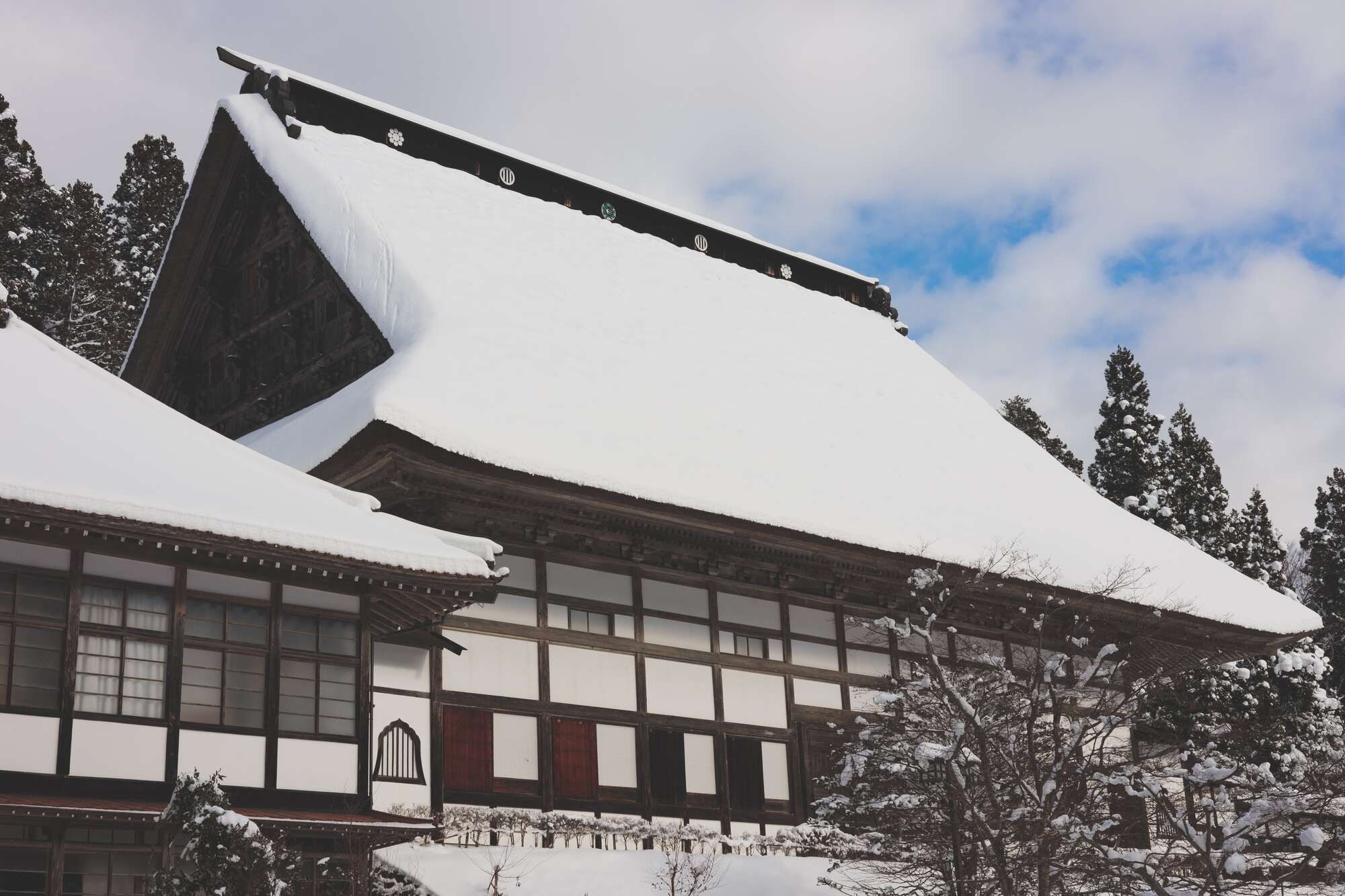 Traditional Japanese building with a snow-covered roof, surrounded by evergreen trees in winter.