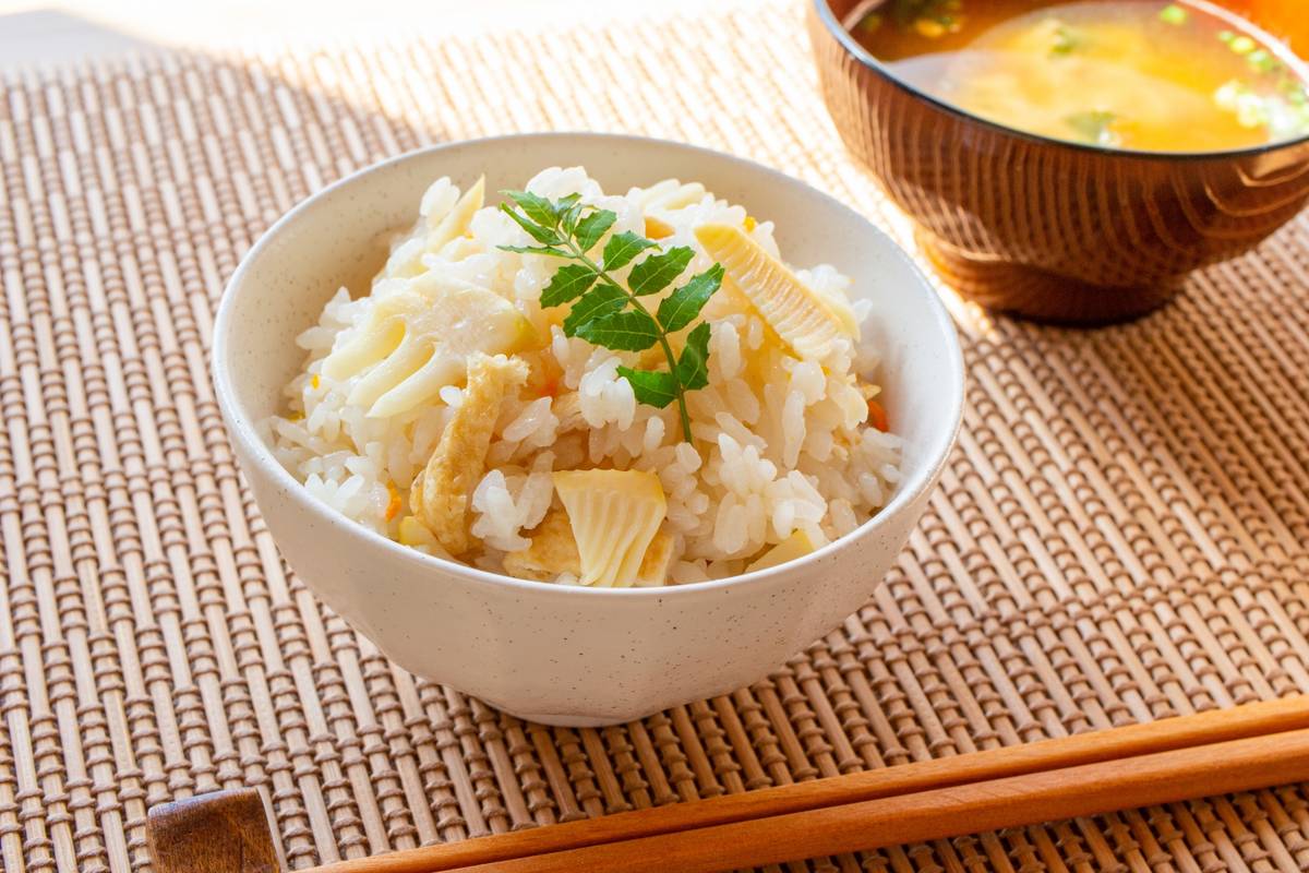 A bowl of bamboo shoot rice, garnished with a leaf, with miso soup and chopsticks on a woven mat.