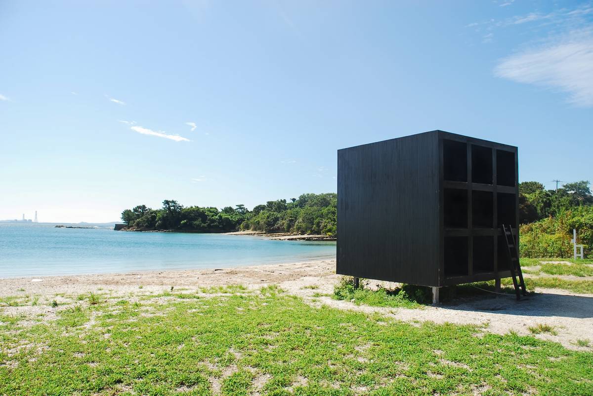 A black cube-shaped structure with open compartments and a ladder stands on a sandy beach next to a calm blue bay and green forested islands.