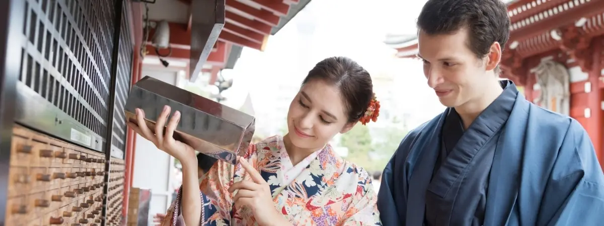 A woman in a kimono shakes a fortune stick box at a Japanese temple while a man watches.