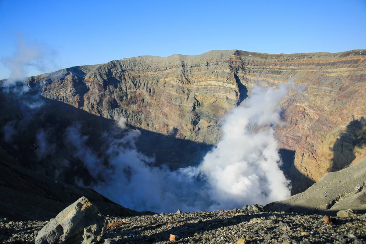 Volcanic crater with stratified rock walls and plumes of steam rising from its base.