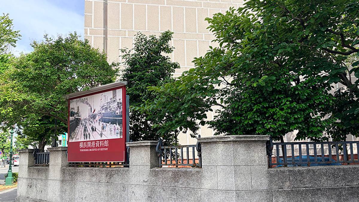 Sign for the Yokohama Archives of History with a historical photo, behind a stone wall with trees.
