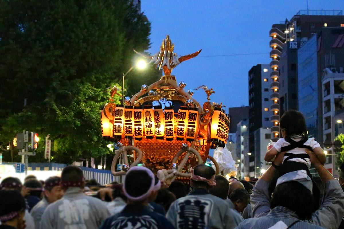 Illuminated Japanese festival float carried by people at dusk, with a child on someone's shoulders in the foreground.