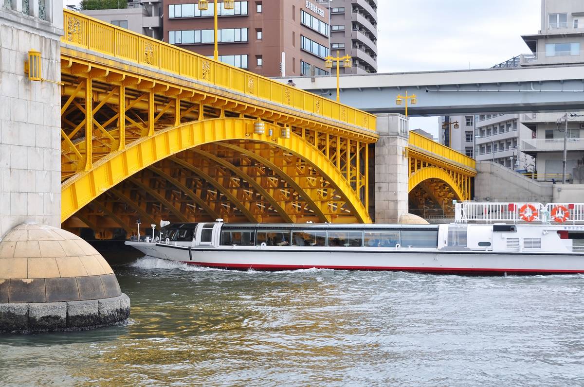A white passenger boat travels on a river under a yellow arched bridge with city buildings in the background.