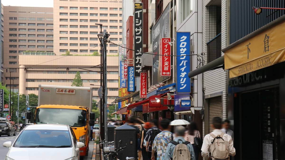 A bustling Japanese city street with blurred pedestrians, cars, and buildings featuring prominent shop signs.
