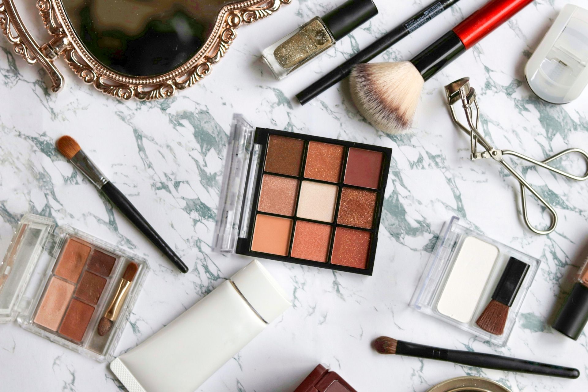 An assortment of makeup products, including eyeshadow palettes, brushes, and a mirror, arranged on a white marble surface.