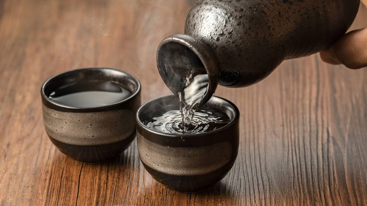 A hand pours steaming liquid from a dark ceramic bottle into a cup, with another cup nearby, on a wooden table.