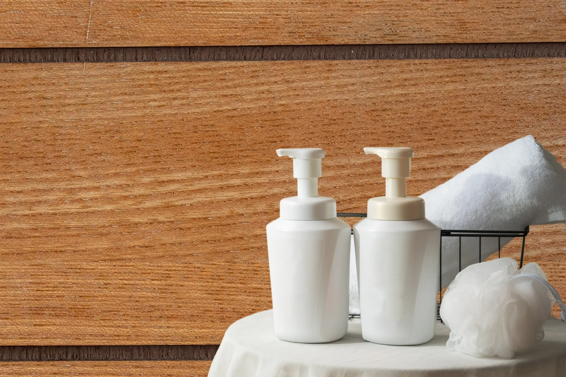 Two white pump bottles, a white towel in a wire basket, and a bath pouf on a white surface with a wooden background.