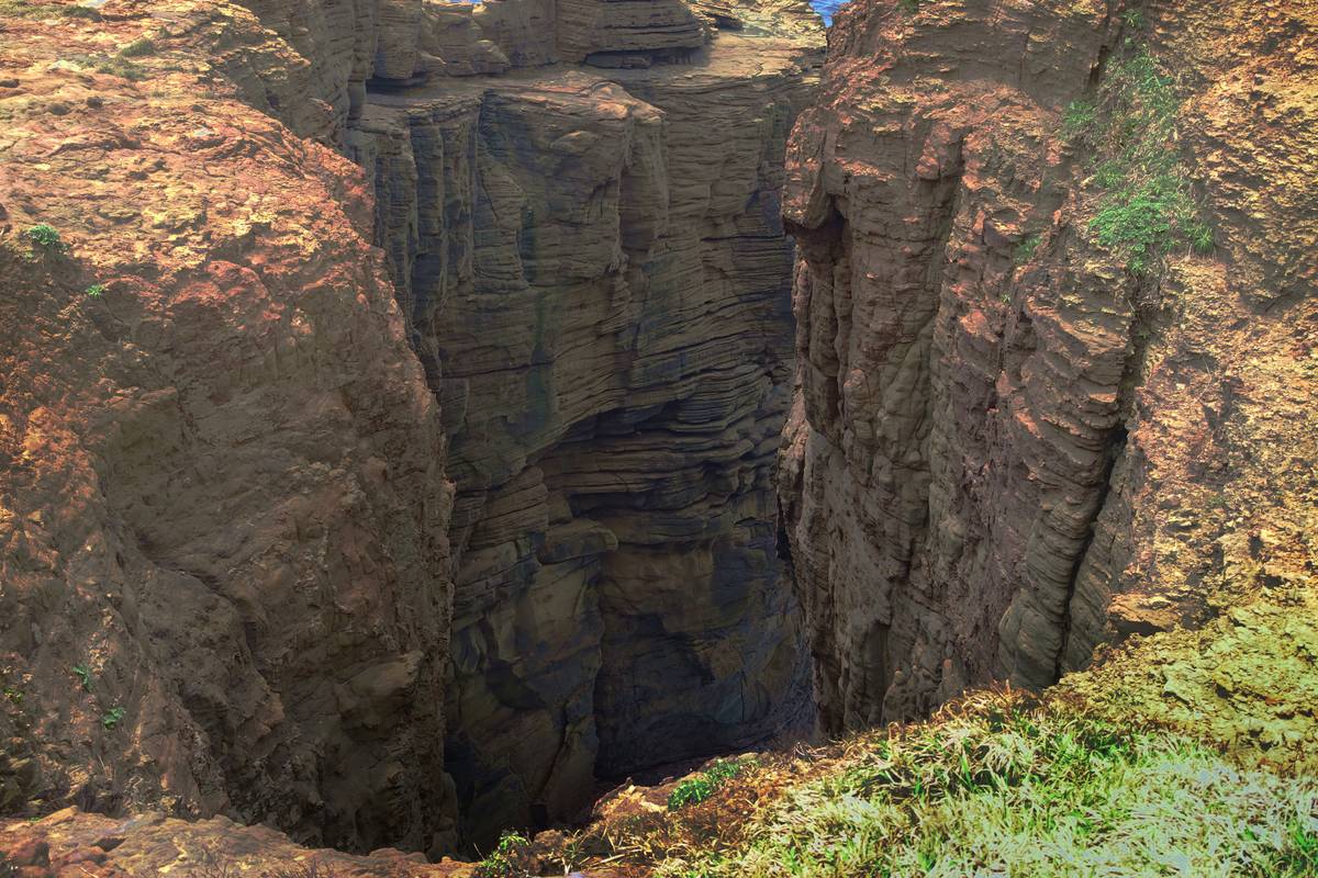 A deep, narrow chasm with steep, layered rock walls and green vegetation at the rim.