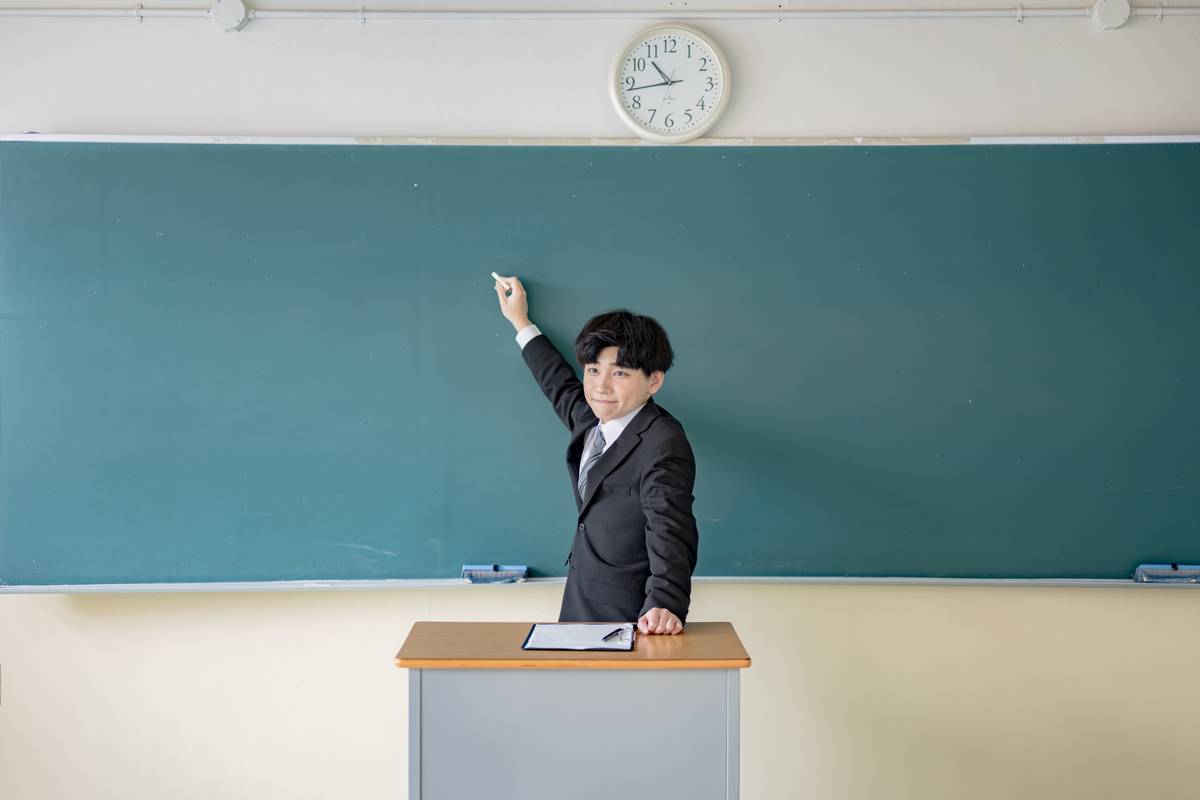 A man in a suit stands at a podium in front of a chalkboard, holding chalk and pointing.