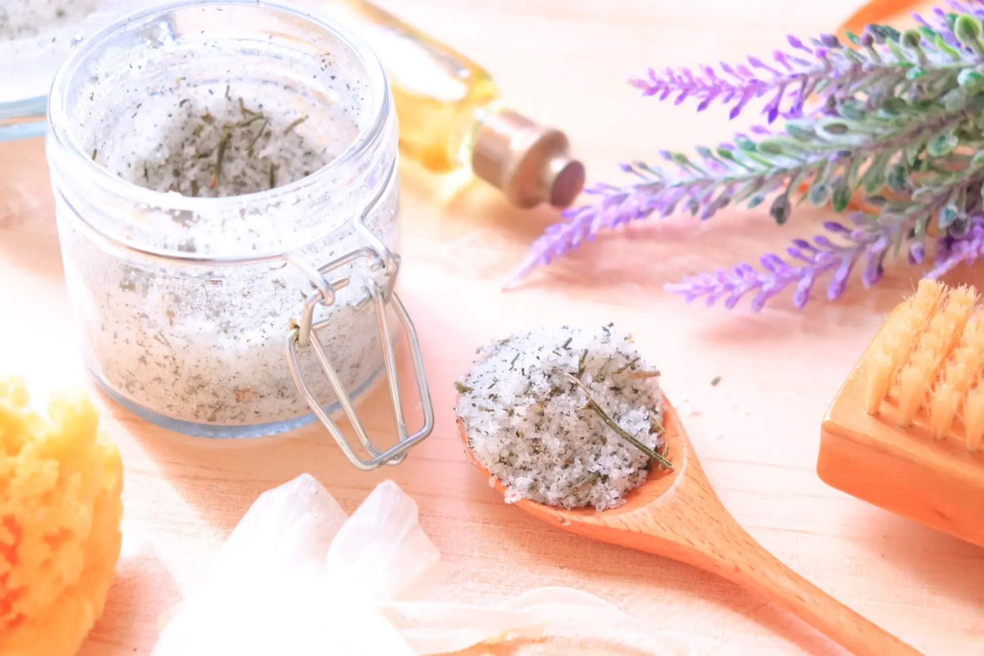 Herbal bath salts in a jar and spoon, with a bottle of oil, lavender, and a brush.