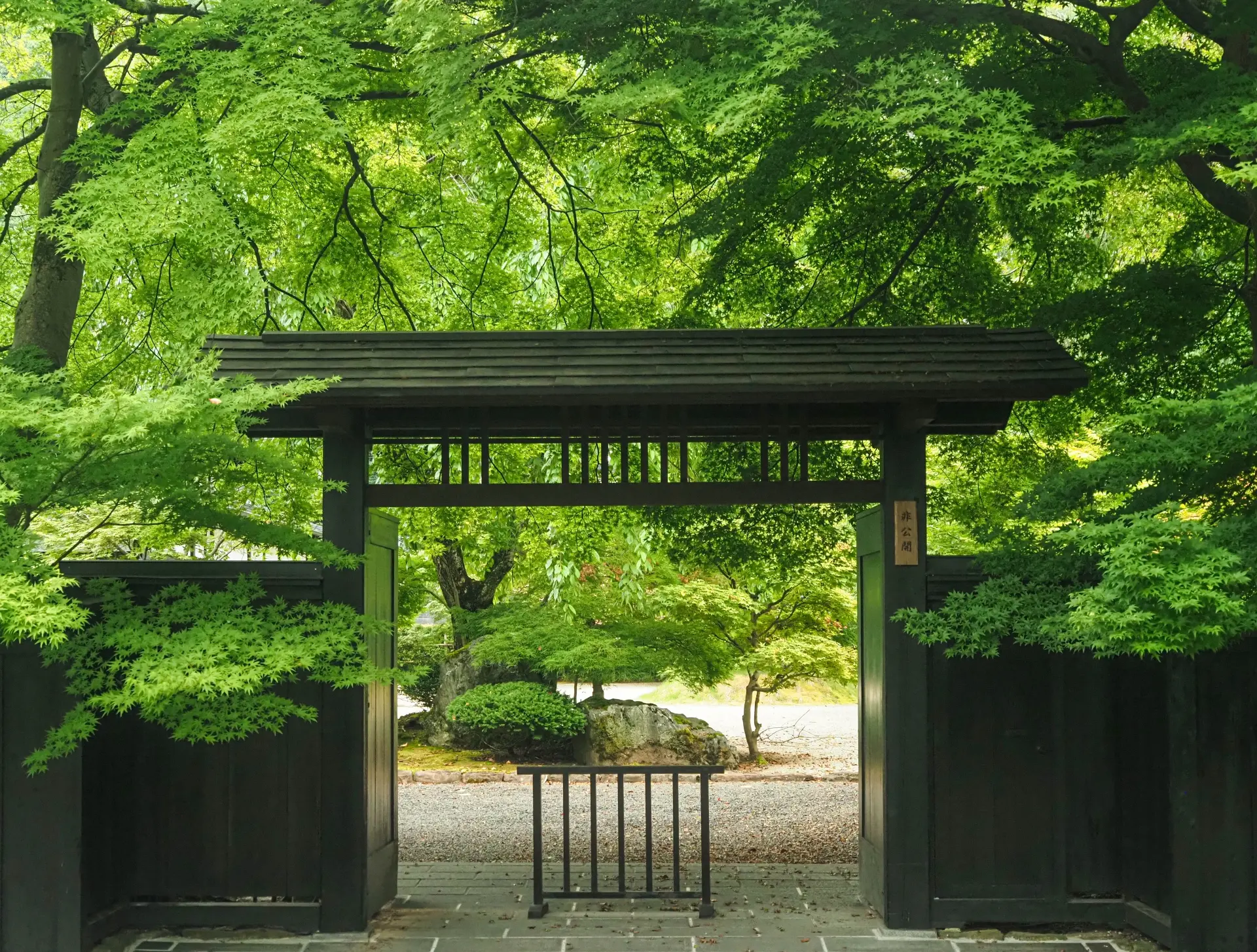 A dark traditional Japanese gate framed by vibrant green foliage, leading to a garden path.