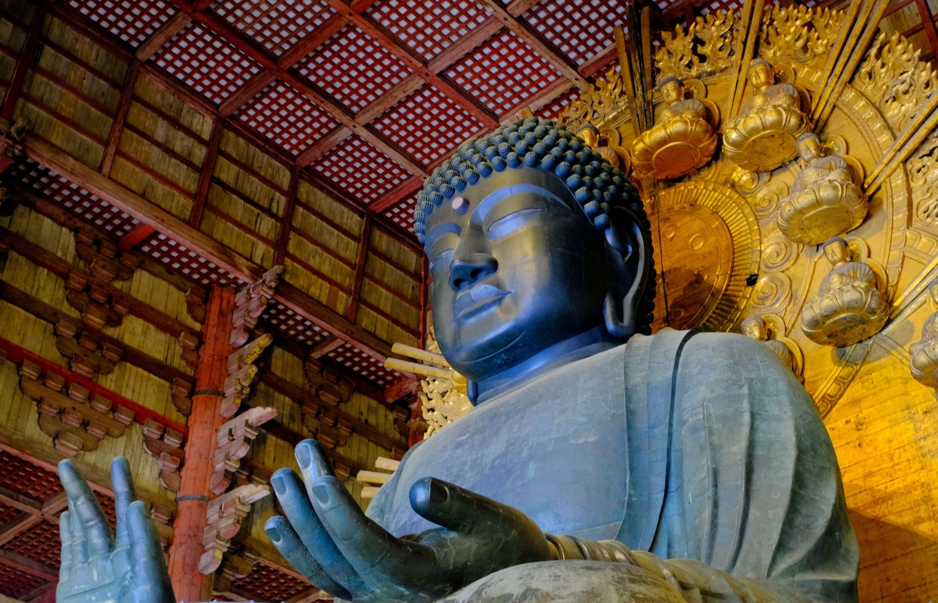 A large, dark Buddha statue with hands raised, in front of a golden halo and ornate wooden temple ceiling.