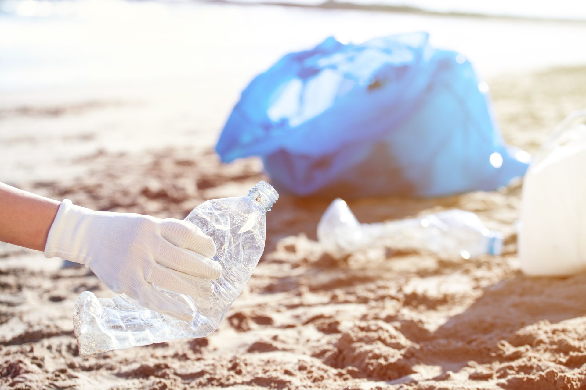 A gloved hand picks up a crushed plastic bottle from a sandy beach, with a blue trash bag in the background.