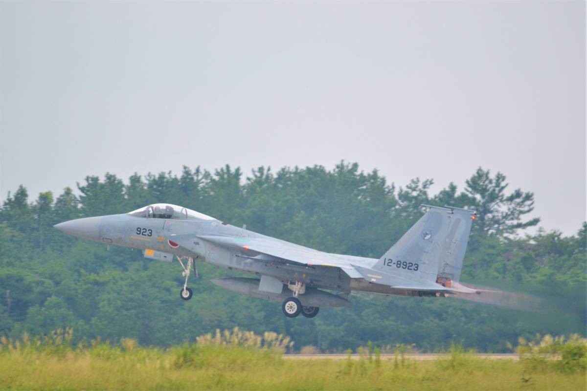 A gray F-15 fighter jet takes off from a runway, with visible exhaust trails and trees in the background.