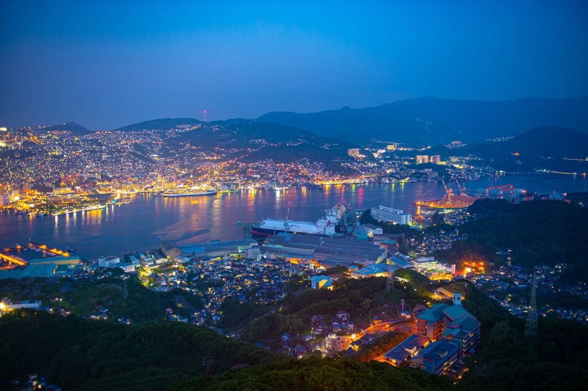 Nighttime panoramic view of a brightly lit port city with lights reflecting on a bay and surrounding hills.