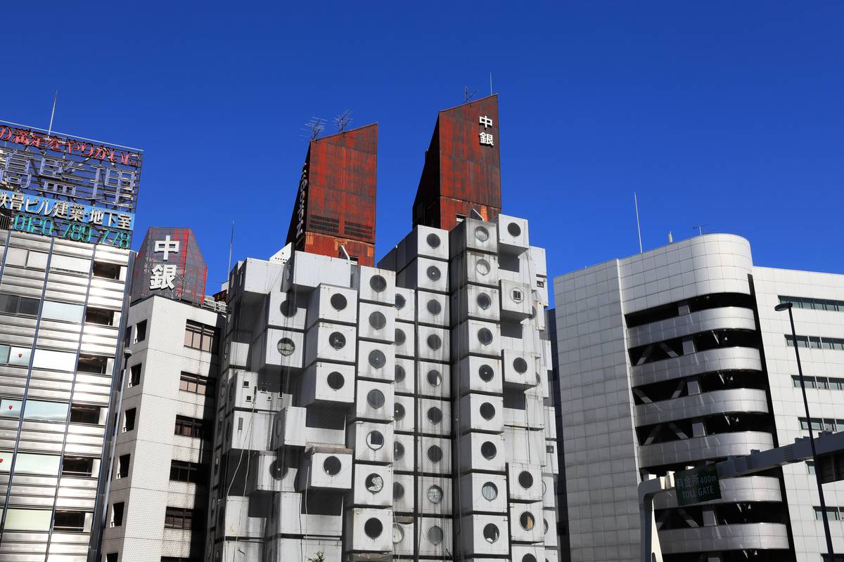 The Nakagin Capsule Tower, a modular building made of square capsules with circular windows, stands among other modern buildings against a bright blue sky.