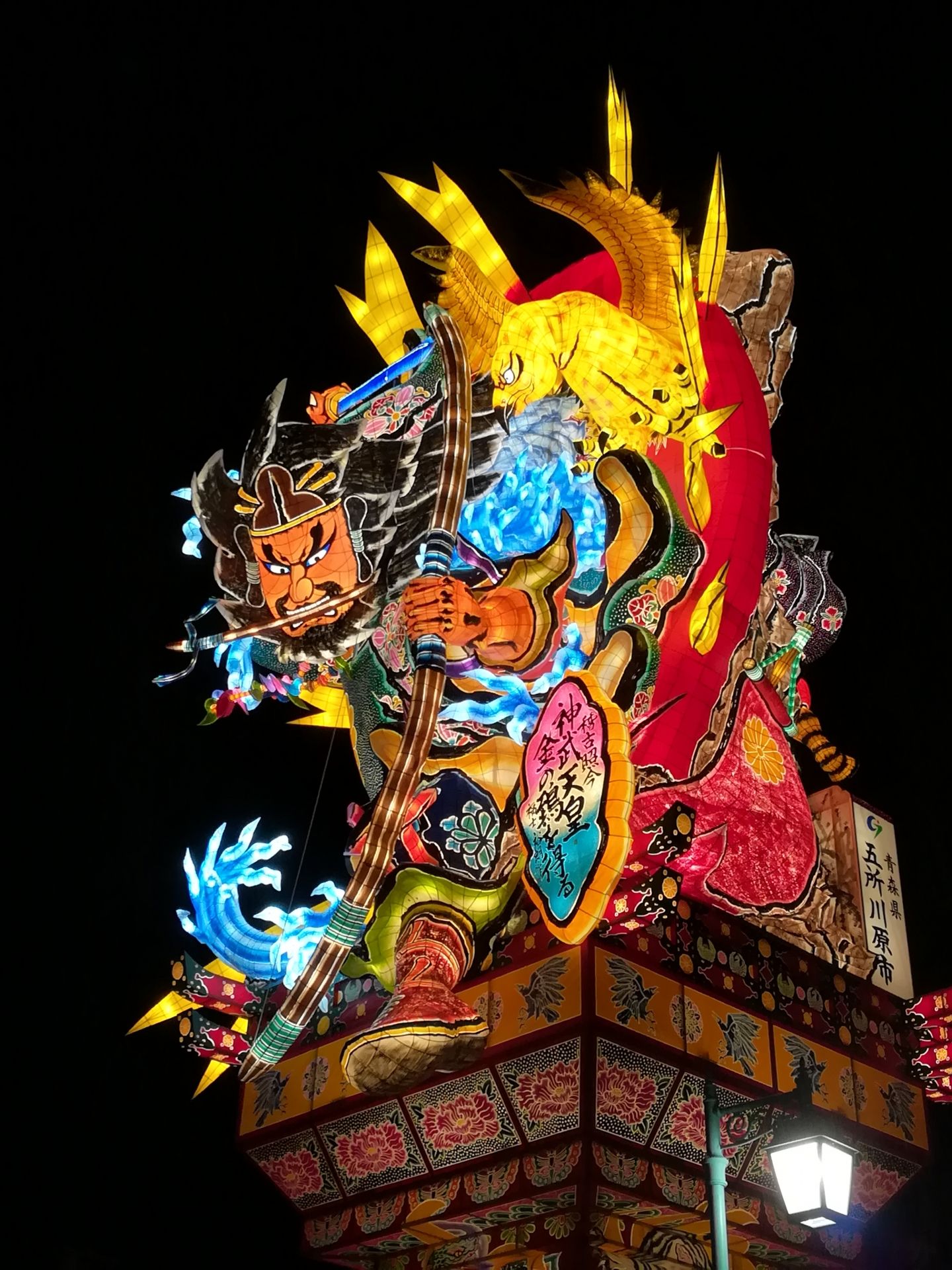 A large, colorful, illuminated Japanese festival float depicting a warrior with a bow against a dark night sky.