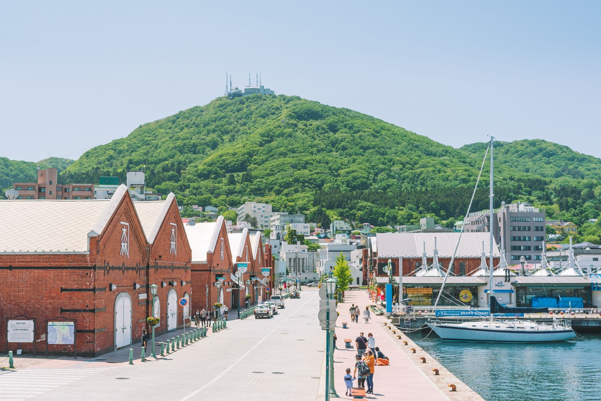 Red brick warehouses line a waterfront street with sailboats docked at a pier, backed by a lush green mountain.