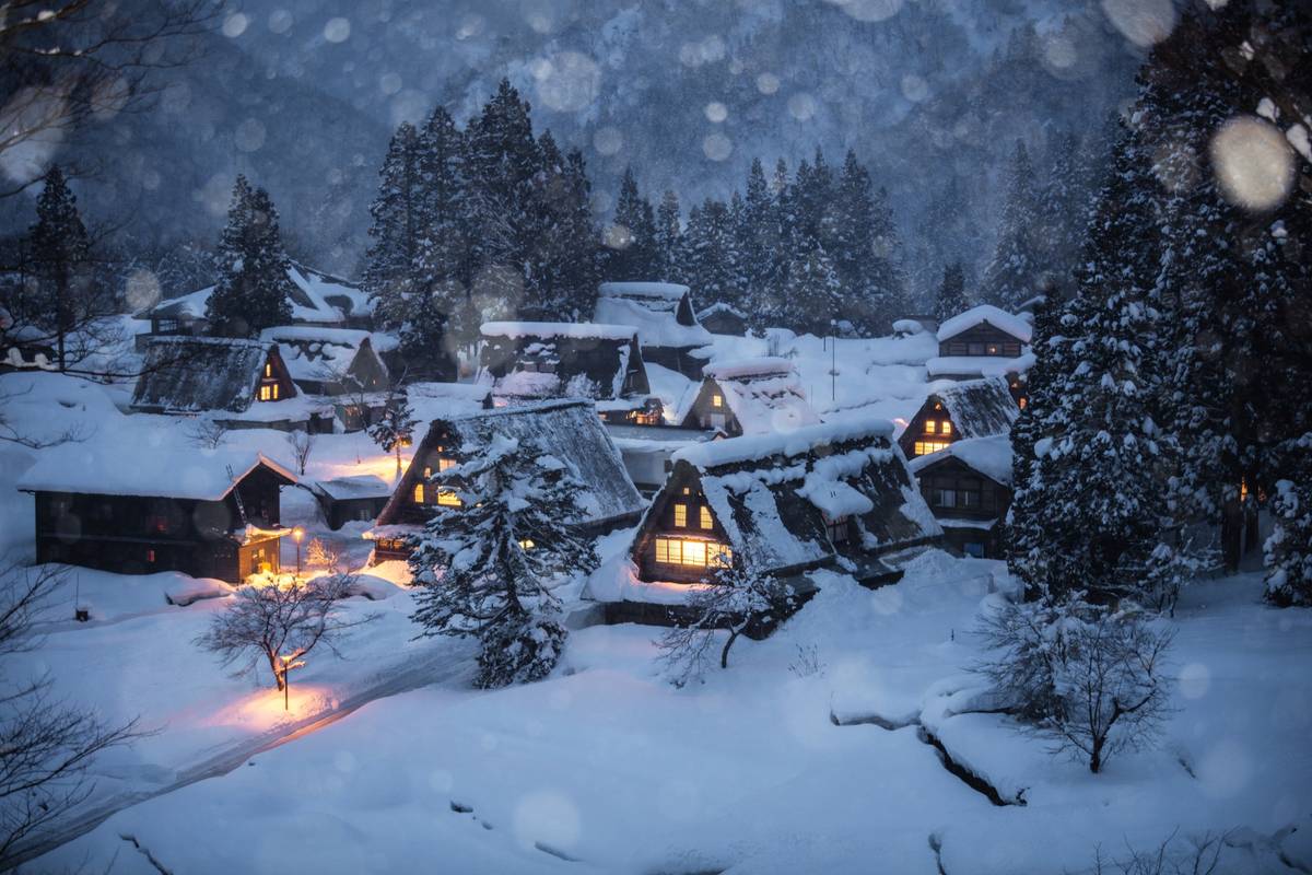 A traditional village in winter, with snow-covered houses glowing with warm light amidst falling snow.