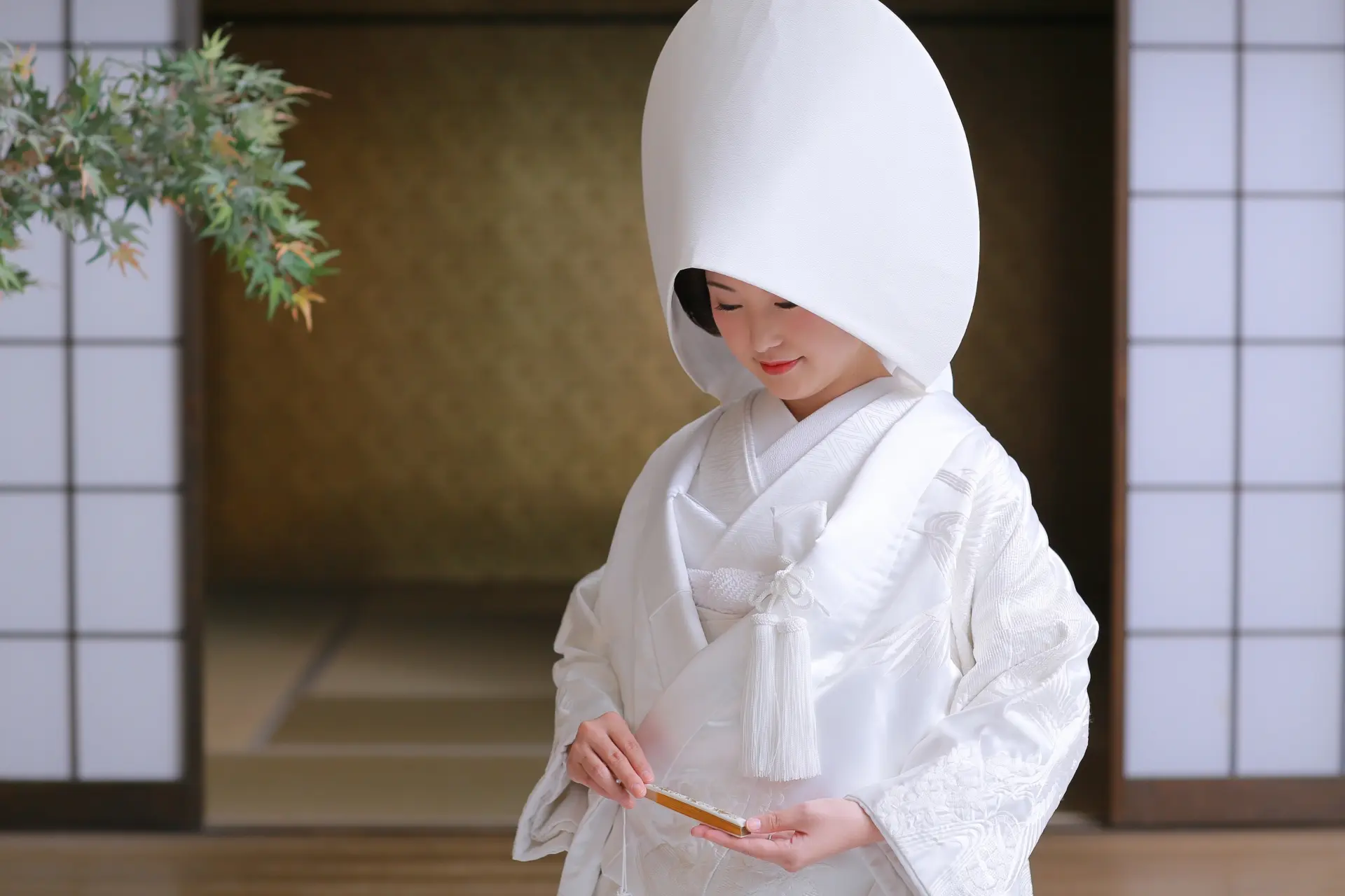 Japanese bride in white kimono and large white headpiece, holding a golden fan.
