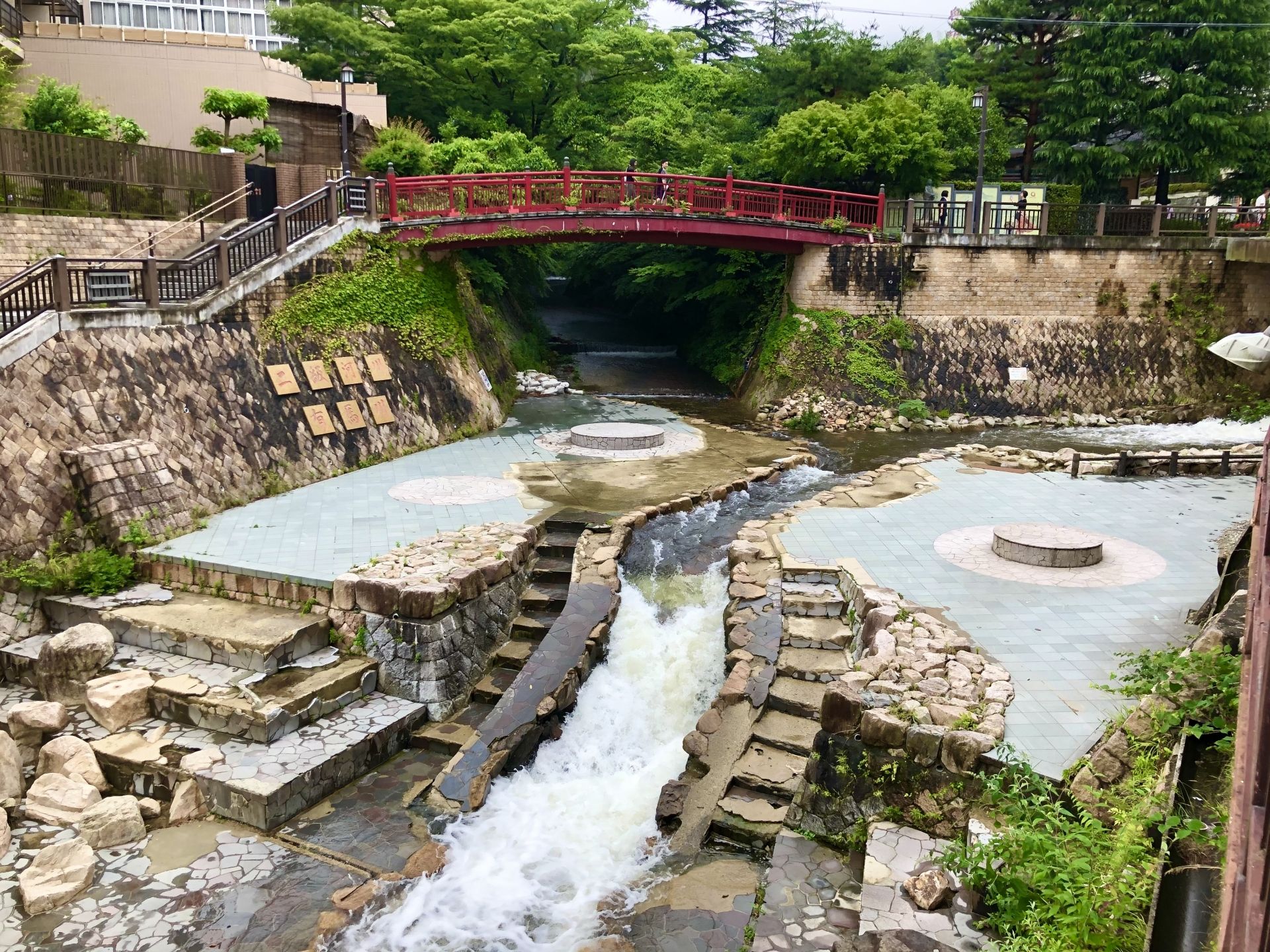 A landscaped stream with cascading water, paved foot bath areas, and a red arched bridge overhead.
