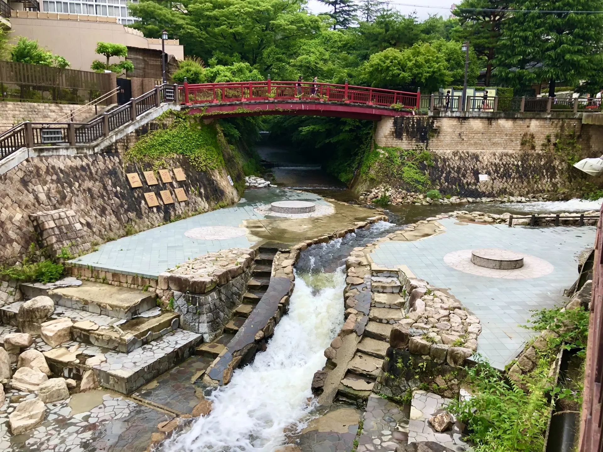 A landscaped stream with cascading water, paved foot bath areas, and a red arched bridge overhead.