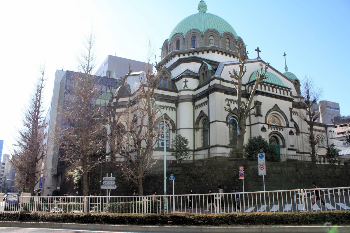 A large, ornate white cathedral with a prominent green dome and crosses, surrounded by bare trees and modern city buildings, with a street in the foreground.