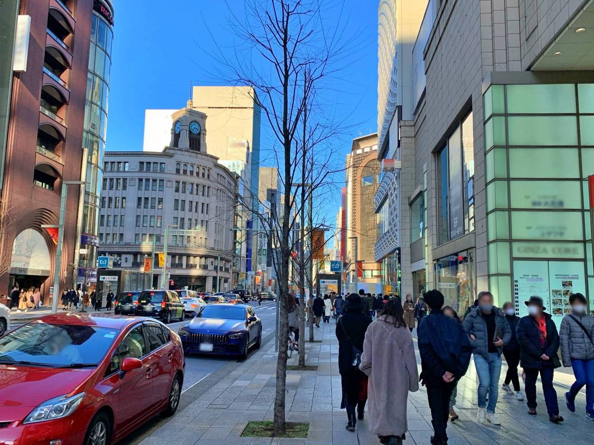 A sunny city street with pedestrians, traffic, tall buildings, and a prominent clock tower under a clear blue sky.