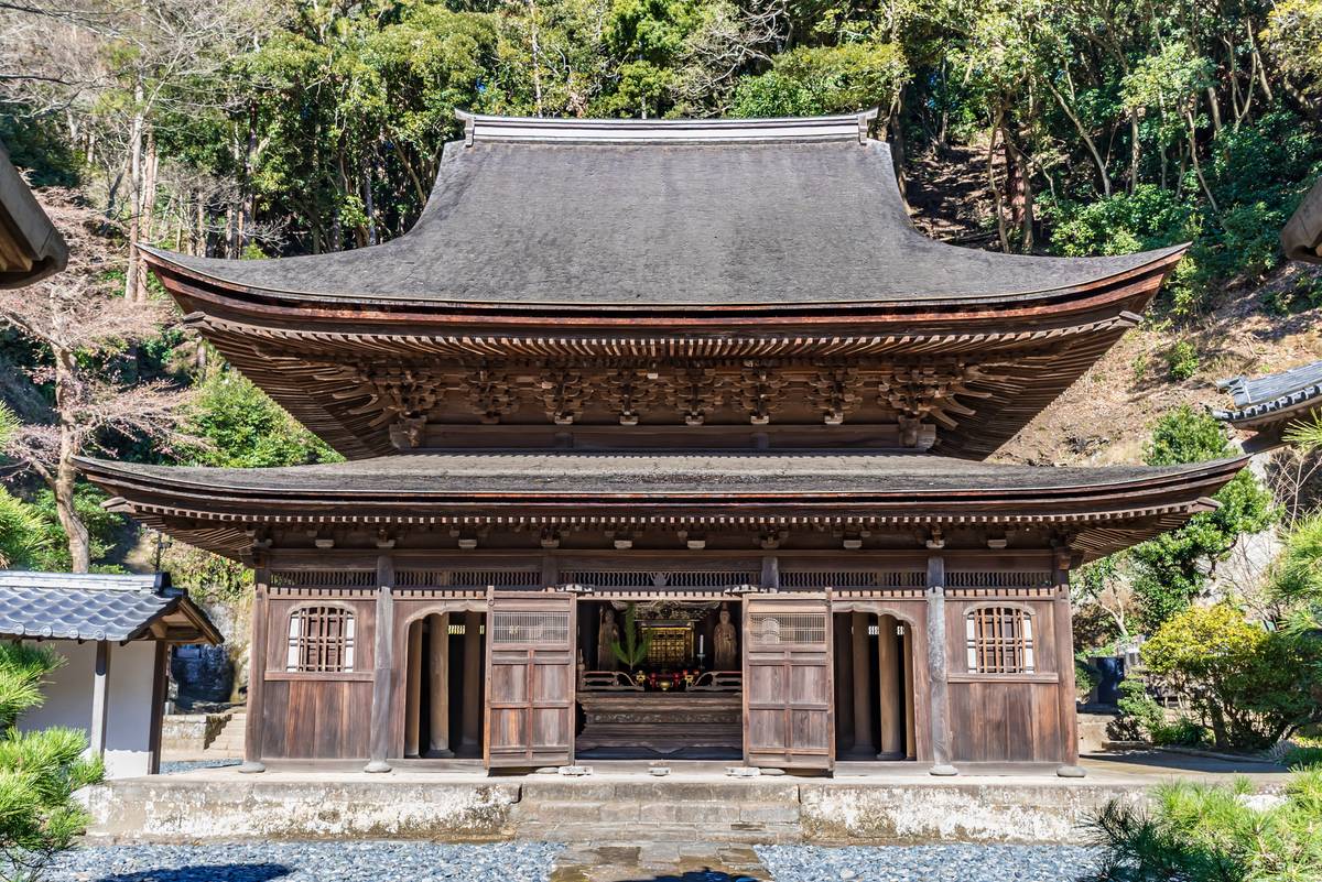 A wooden Japanese temple with a multi-tiered roof and open entrance, set against a forest background.