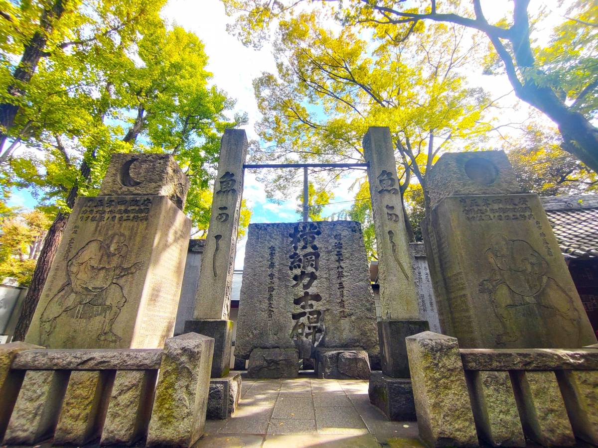 Stone monuments with carved sumo wrestlers and Japanese text beneath bright green and yellow trees.