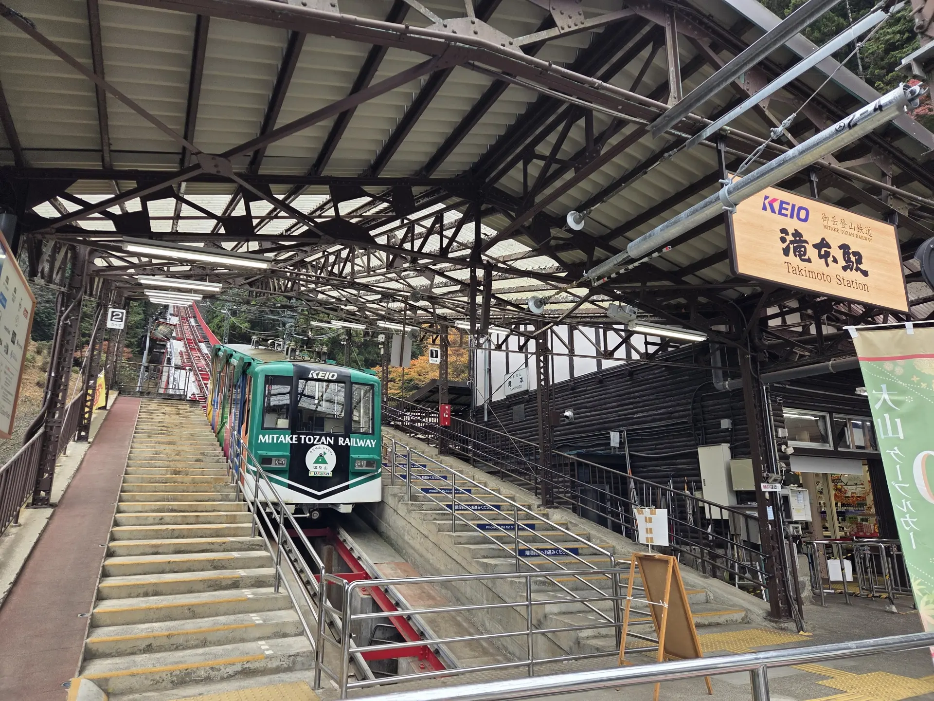 A green and white Mitake Tozan Railway funicular train on an inclined track at Takimoto Station, with stairs alongside.