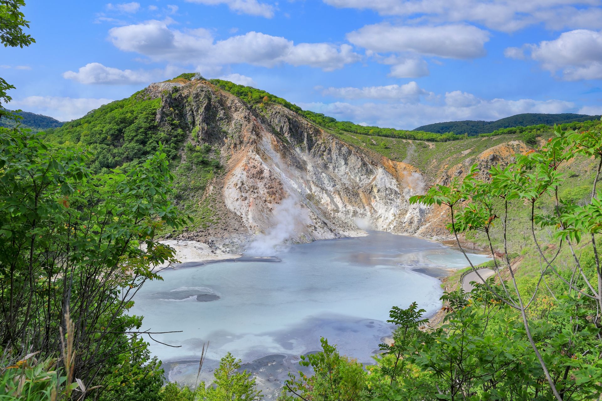 Steaming volcanic crater lake with pale blue water, surrounded by green hills and rocky slopes under a blue sky.