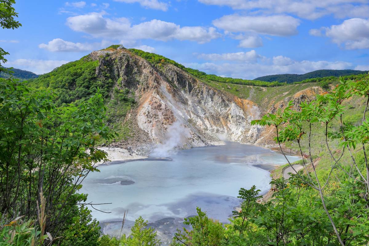 Steaming volcanic crater lake with pale blue water, surrounded by green hills and rocky slopes under a blue sky.