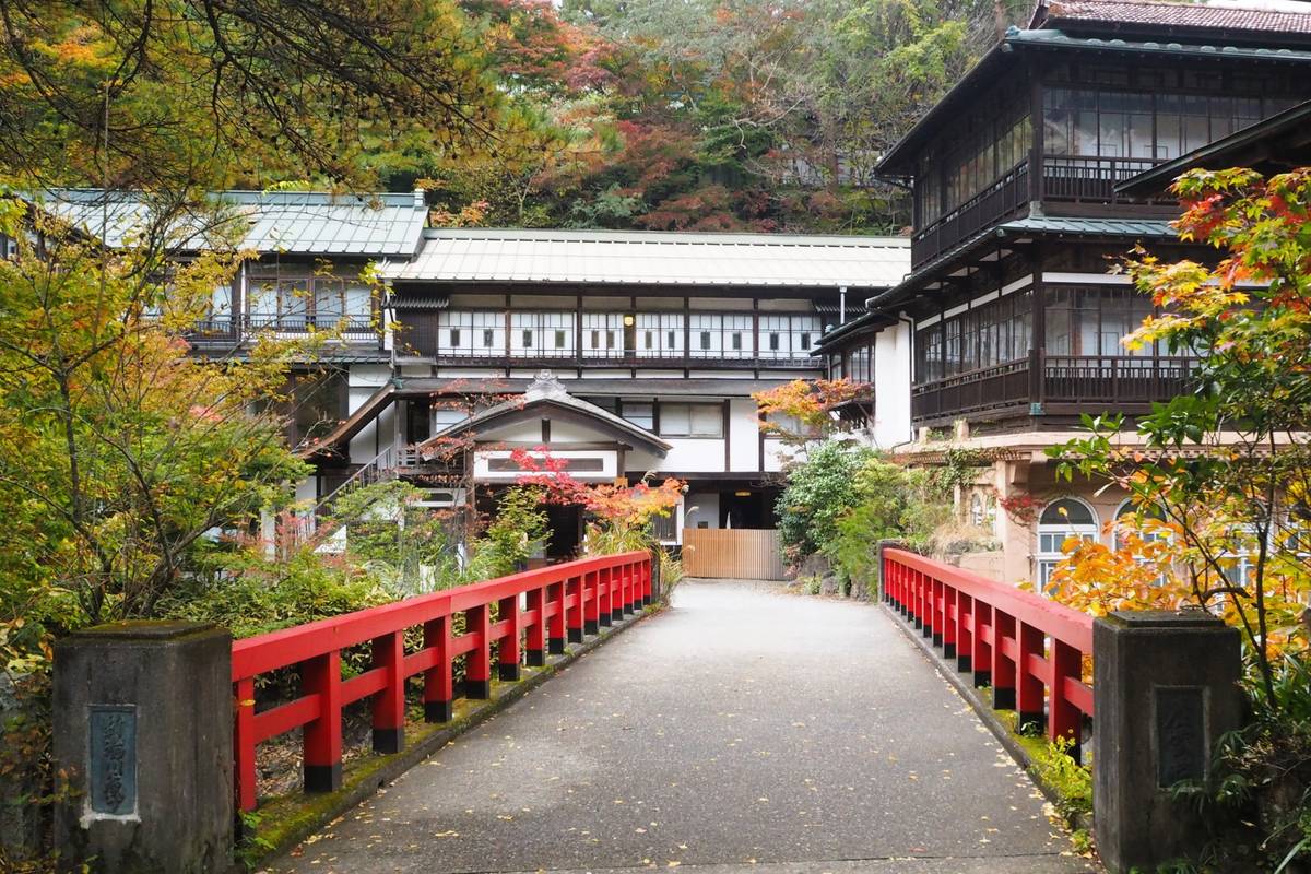 A red bridge leads to traditional Japanese buildings surrounded by vibrant autumn trees.