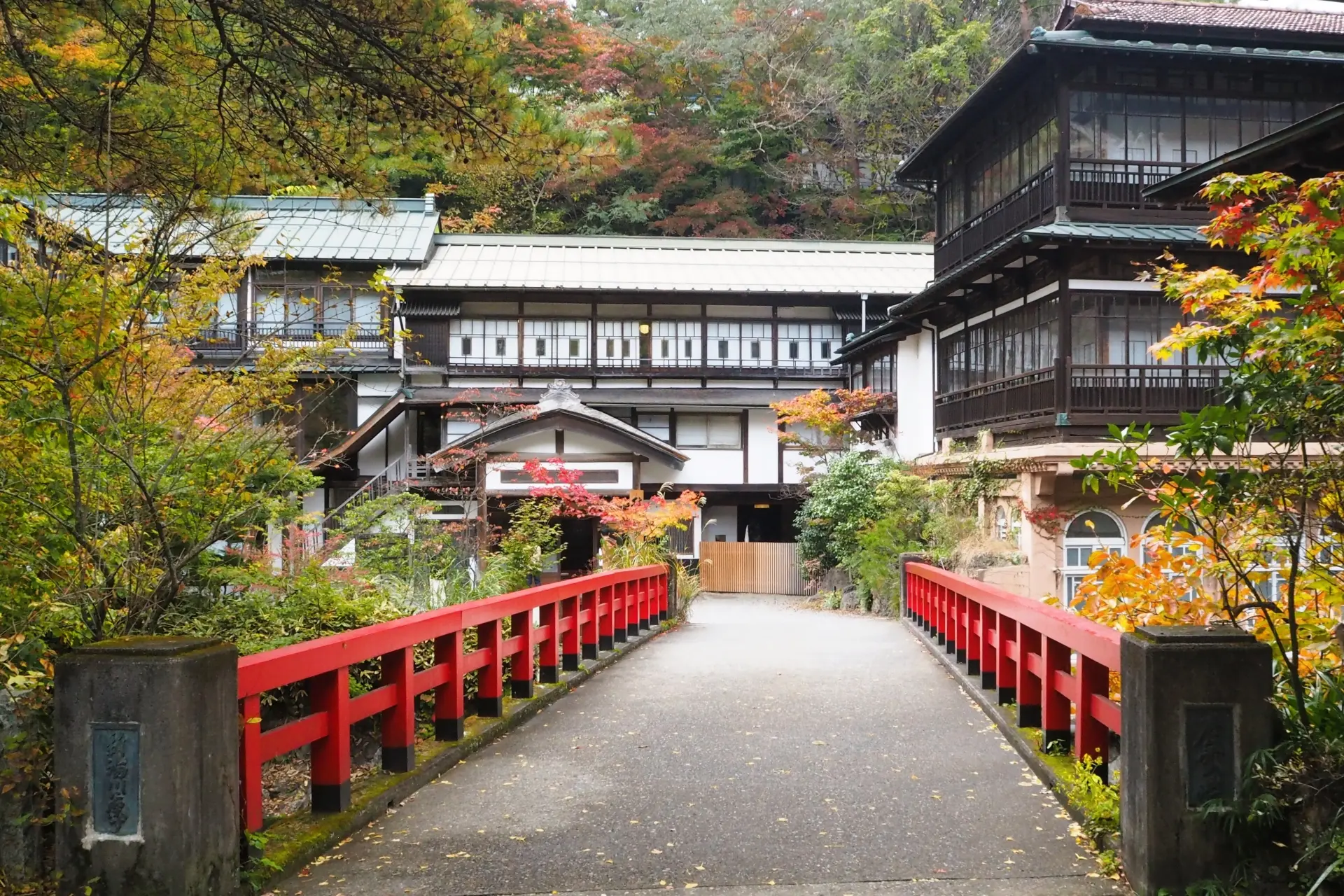 A red bridge leads to traditional Japanese buildings surrounded by vibrant autumn trees.