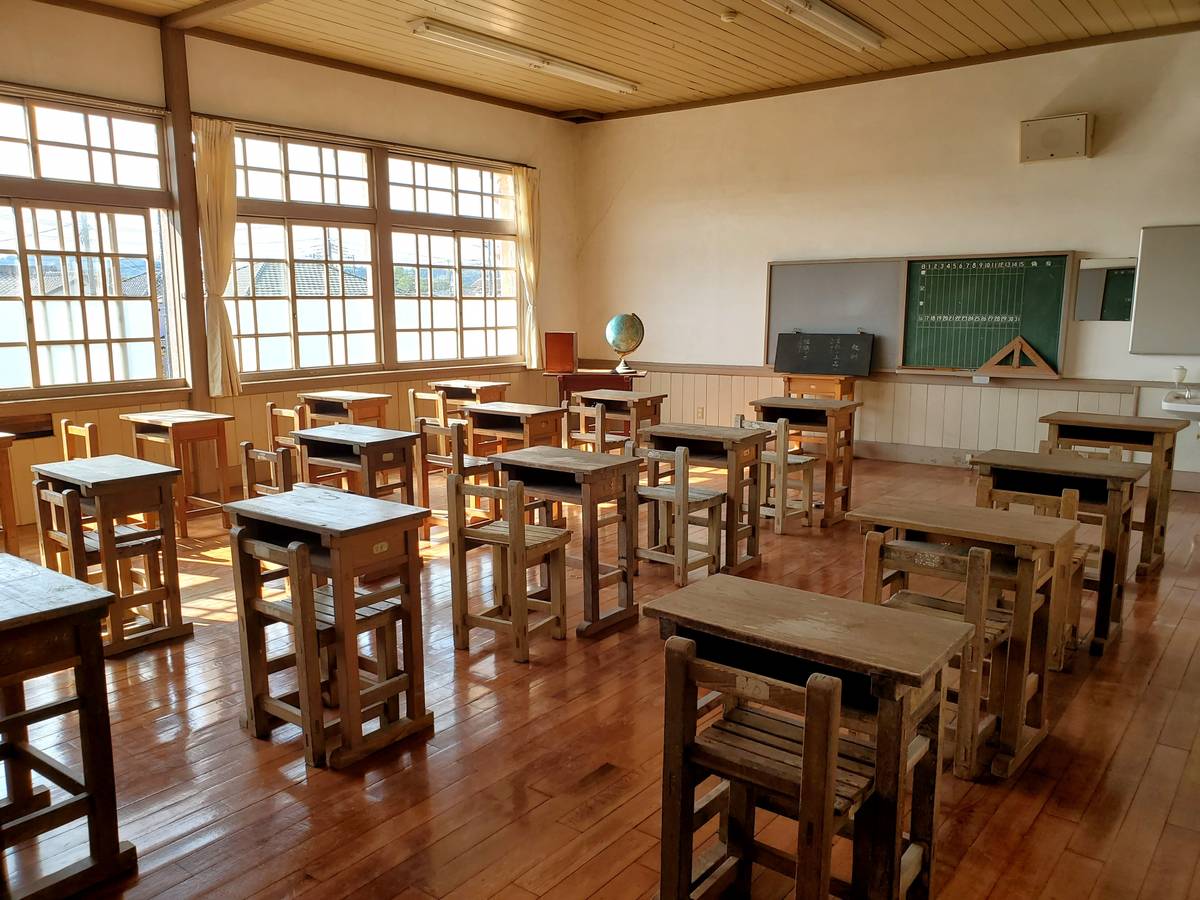 An empty traditional classroom with rows of wooden desks and chairs, large windows, and a blackboard.