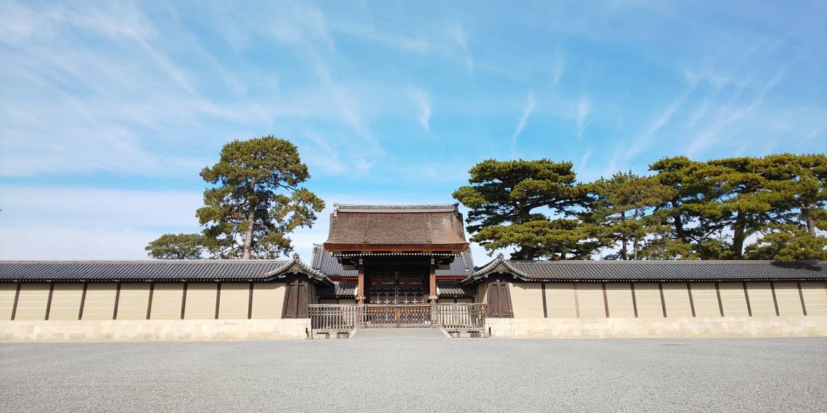 A traditional Japanese gatehouse with tiled walls, flanked by pine trees, under a blue sky over a wide gravel courtyard.