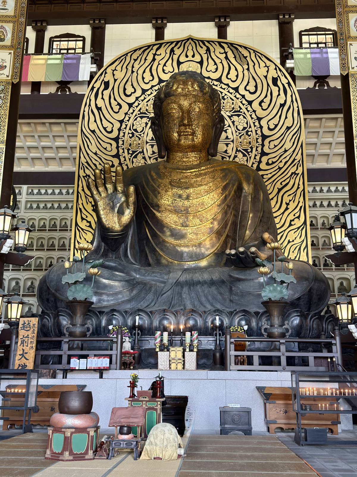 A colossal bronze Buddha with a golden face and elaborate gold halo, seated in a temple with offerings and a person kneeling.
