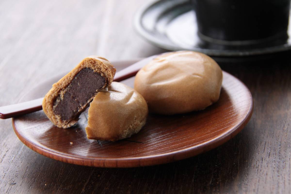 Three light brown manju on a wooden plate, one cut open to reveal a dark red bean paste filling.