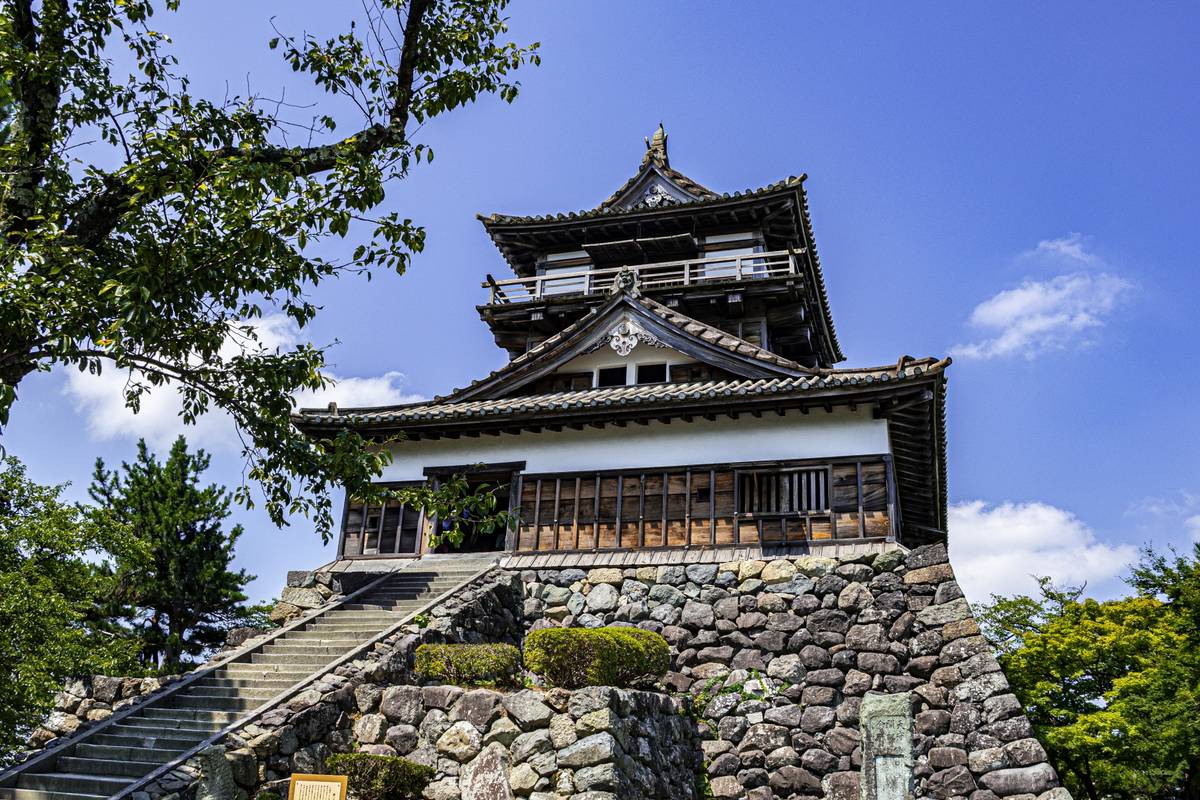 A Japanese castle with a dark multi-tiered roof and white walls, atop a stone foundation, with stairs leading up, under a bright blue sky.