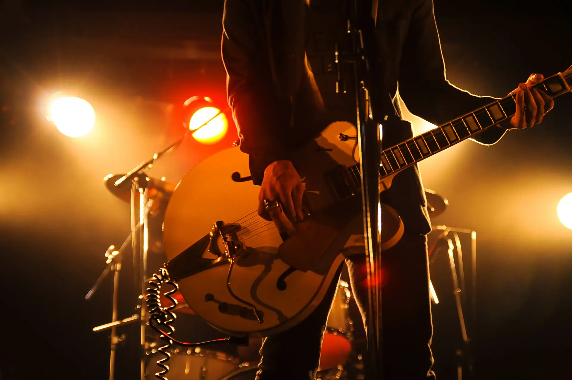 A musician plays an electric guitar on stage under warm, bright spotlights.