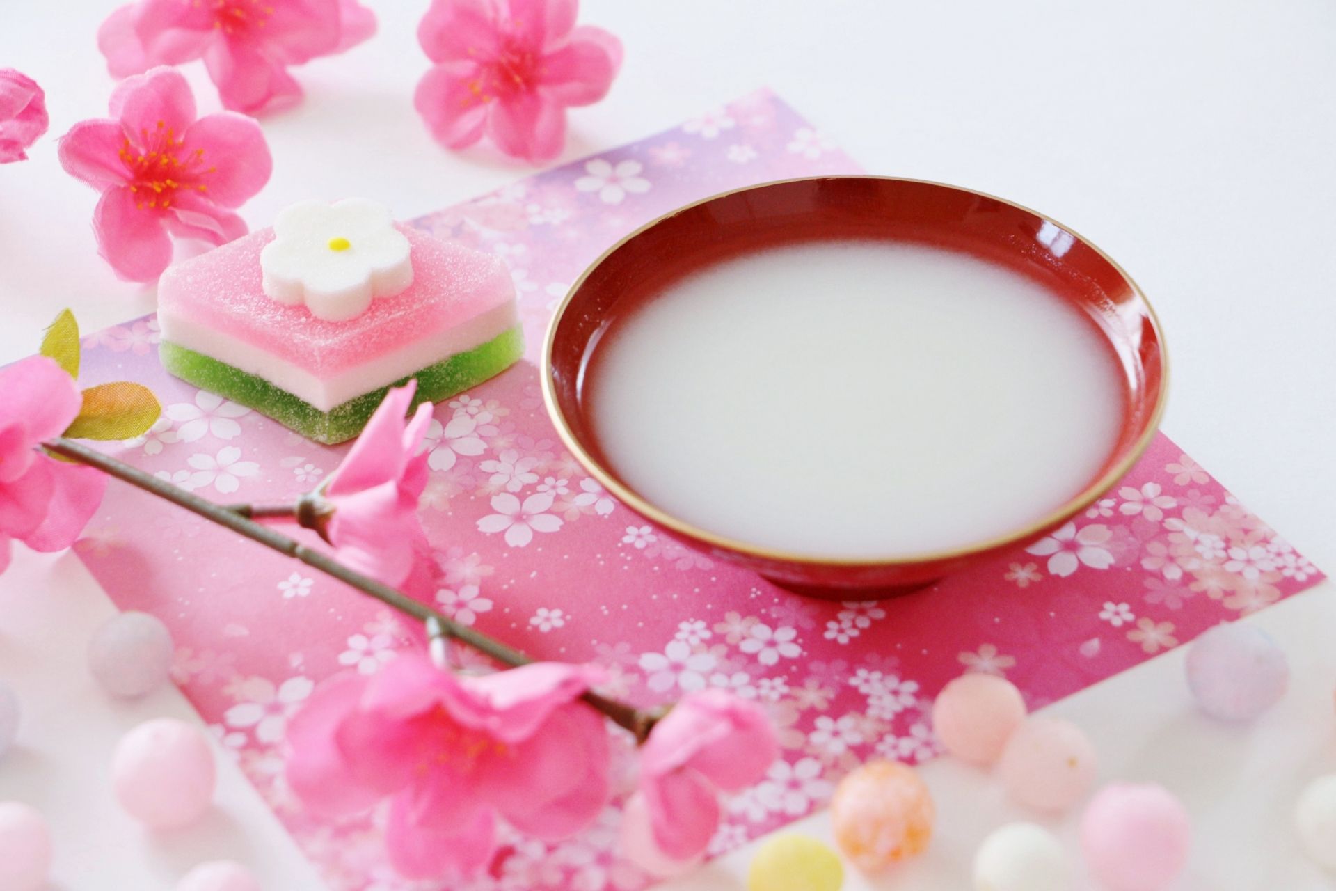 Japanese layered sweet, a bowl of white drink, and pink cherry blossoms on a patterned mat.