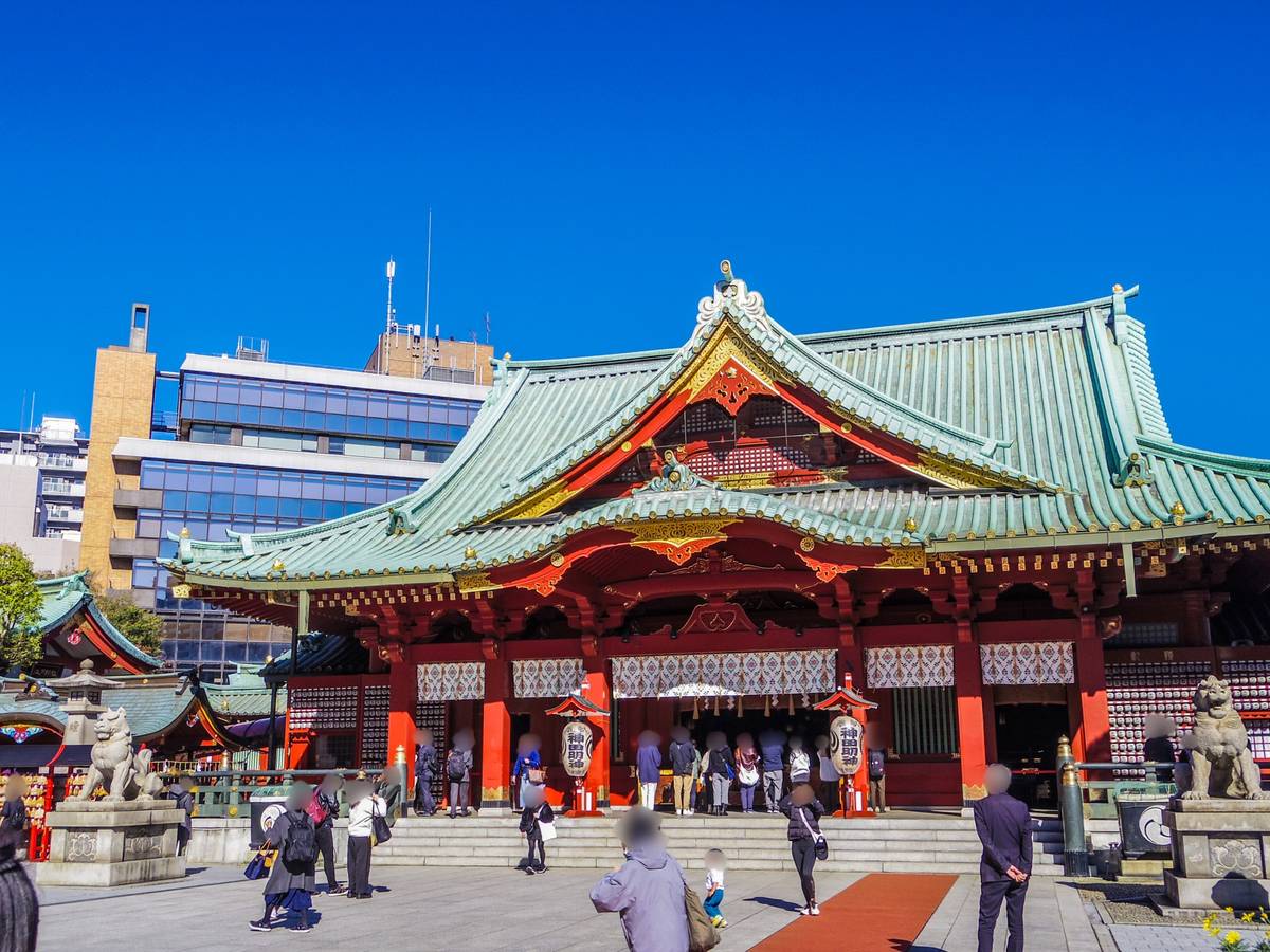 A bright red and gold Japanese shrine with a green roof, people in front, and modern buildings in the background.