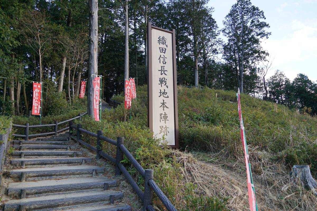 Stone steps lead up a hill, past a large sign in Japanese marking Oda Nobunaga's battlefield headquarters and several red banners.