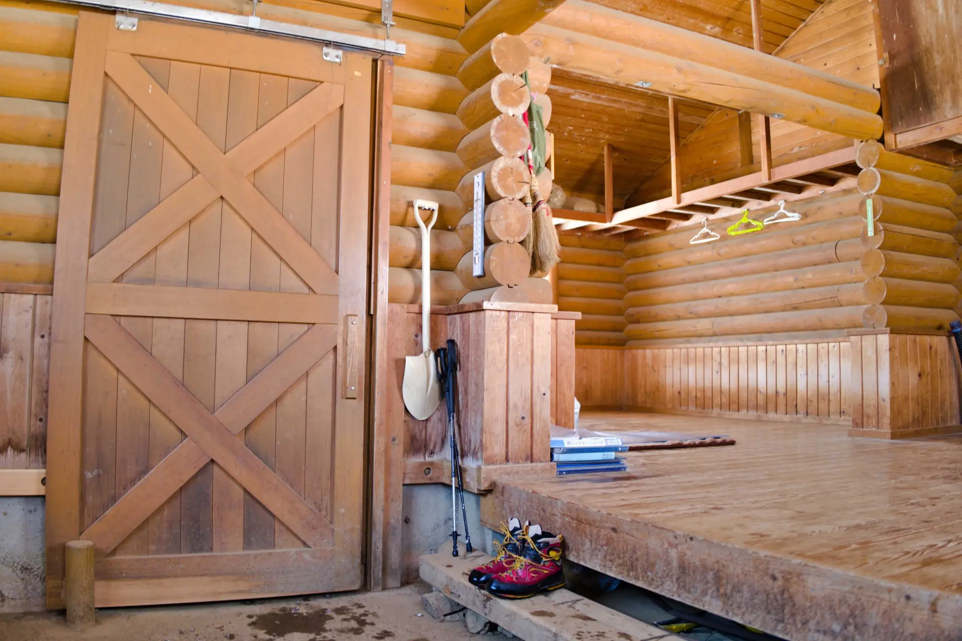 Rustic log cabin interior with a large sliding barn door, a raised wooden platform, and outdoor gear.