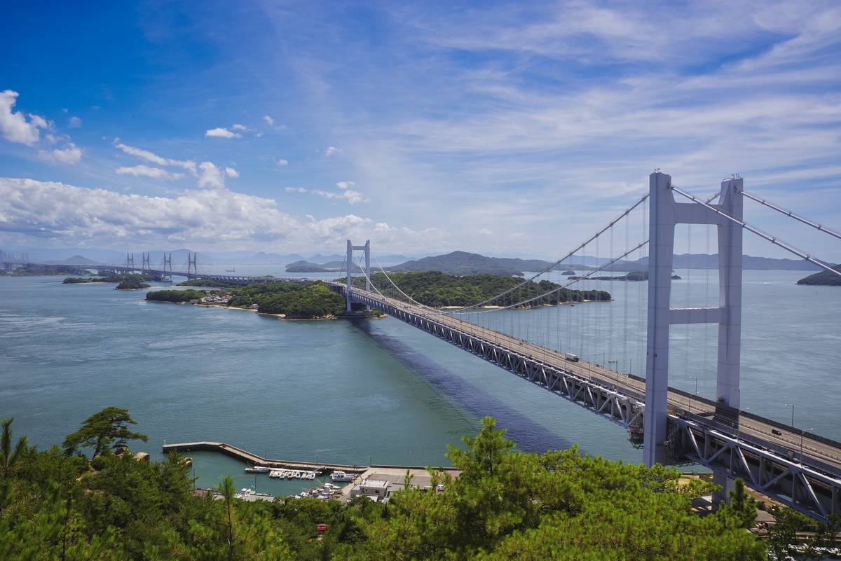 A large suspension bridge crosses an island-dotted sea, with other bridges in the distance under a blue sky.