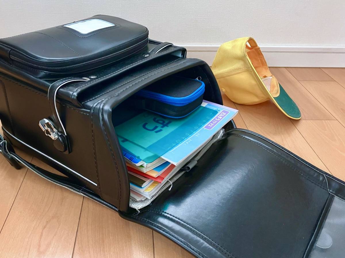 An open black randoseru backpack filled with books and a pencil case, with a yellow cap nearby on a wooden floor.