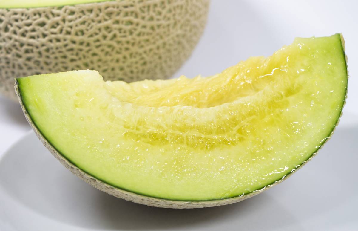 A slice of green-skinned melon with pale yellow flesh, beside a blurred whole melon.